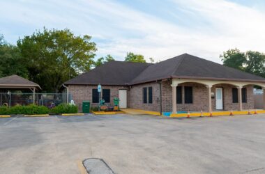Luv-N-Care Learning Center exterior view showcasing a brick building with a sloped roof, surrounded by greenery and a parking area, emphasizing a safe and welcoming environment for children.